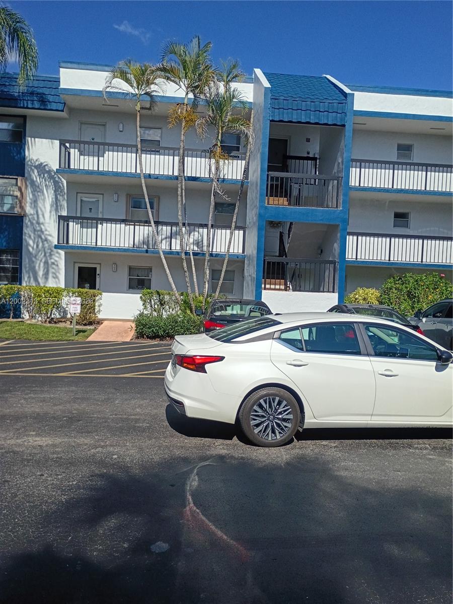 A three-story apartment building with blue trim and balconies stands behind a parking lot — 3040 Holiday Springs Blvd # 110, Margate FL 33063, MARGATE, FL