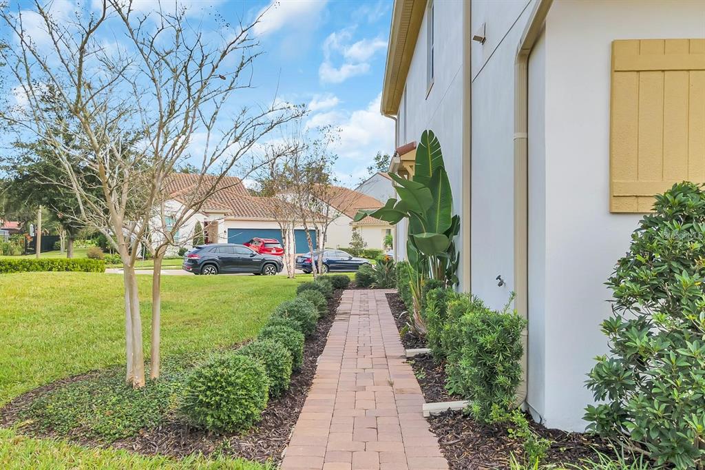 A paved walkway runs alongside a house exterior with manicured shrubs and a grassy lawn — 2795 BOLZANO DRIVE, APOPKA, FL