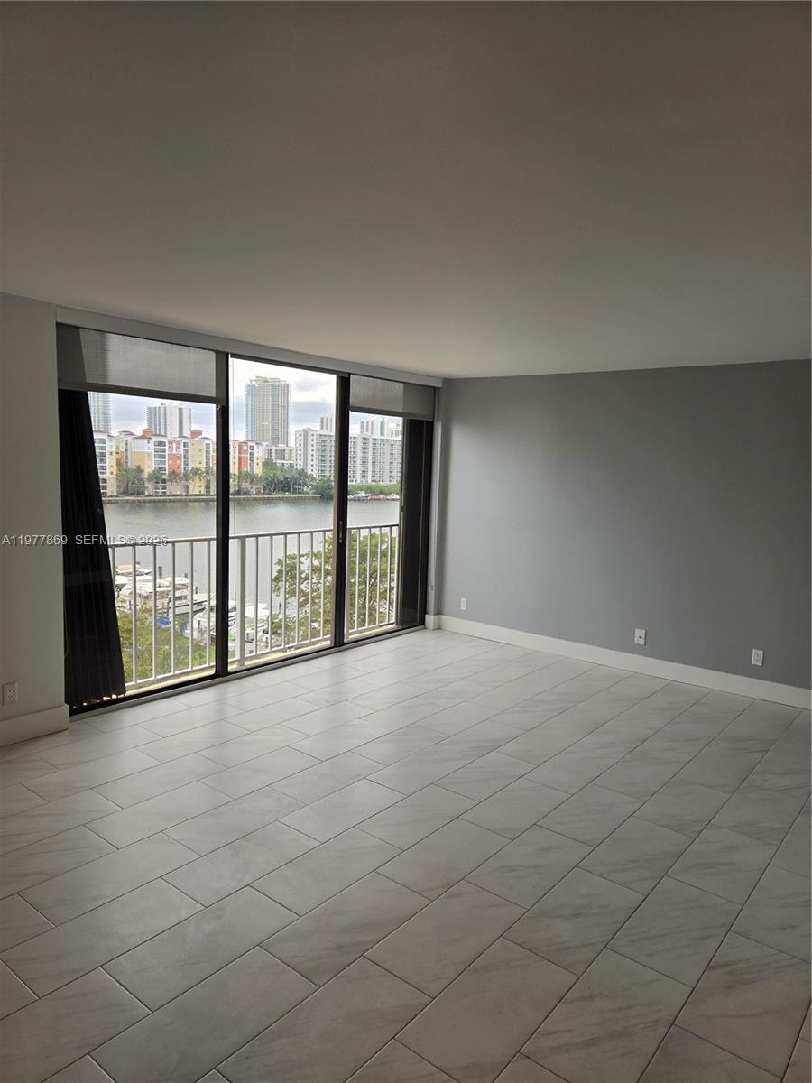 An empty living room features light-colored tile flooring, gray walls, and a balcony overlooking water — SUNNY ISLES BEACH, FL