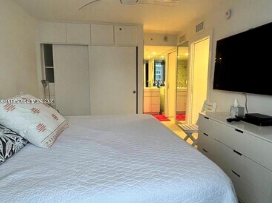 A bedroom features a white bed, a dresser with a television, and a doorway leading to a bathroom — 2457 Collins Ave # 1605, Miami Beach FL 33140, MIAMI BEACH, FL