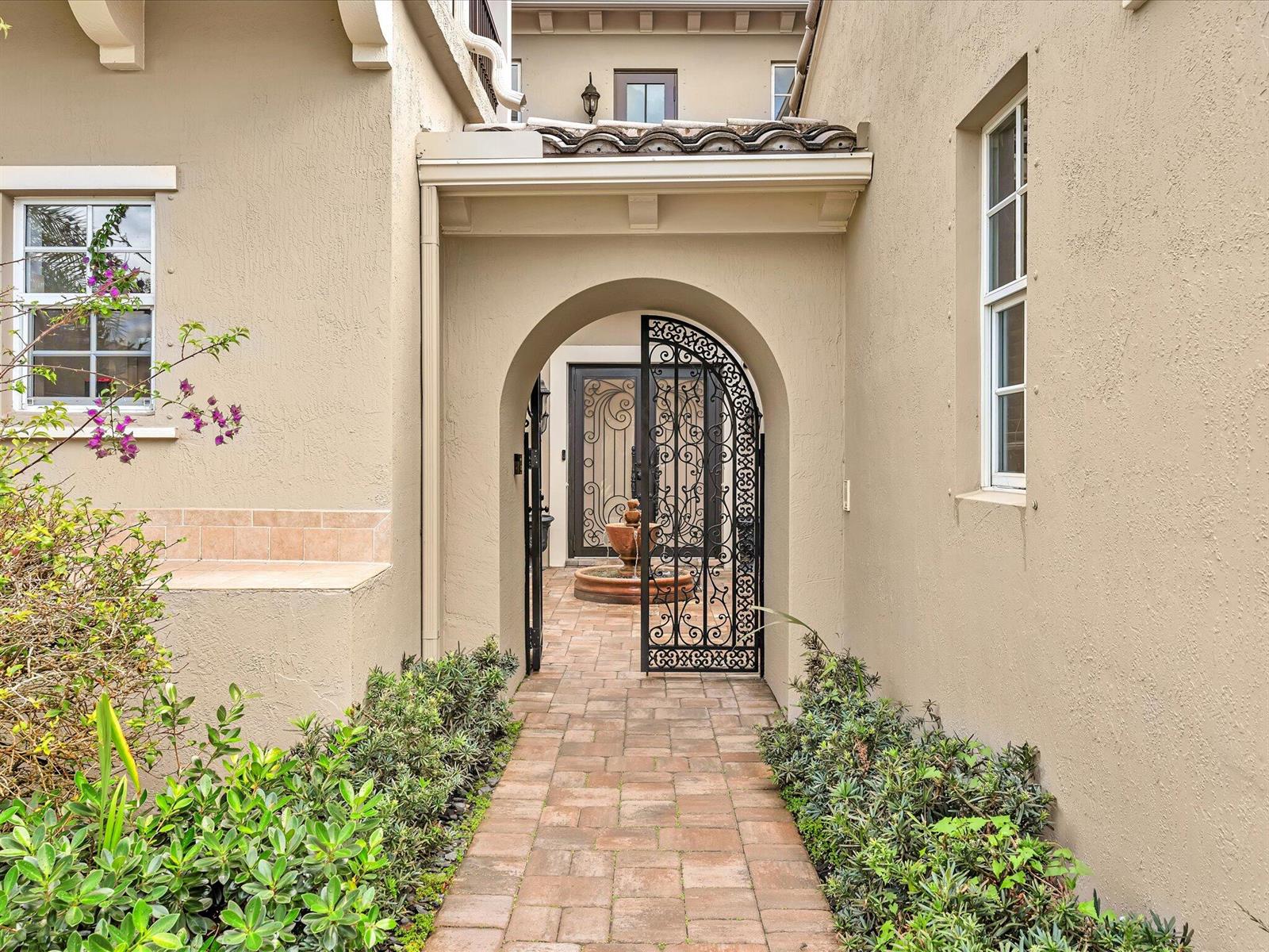 A brick walkway leads through an arched wrought iron gate to a courtyard with a fountain — 10982 NW 81st Manor, Parkland FL 33076, PARKLAND, FL