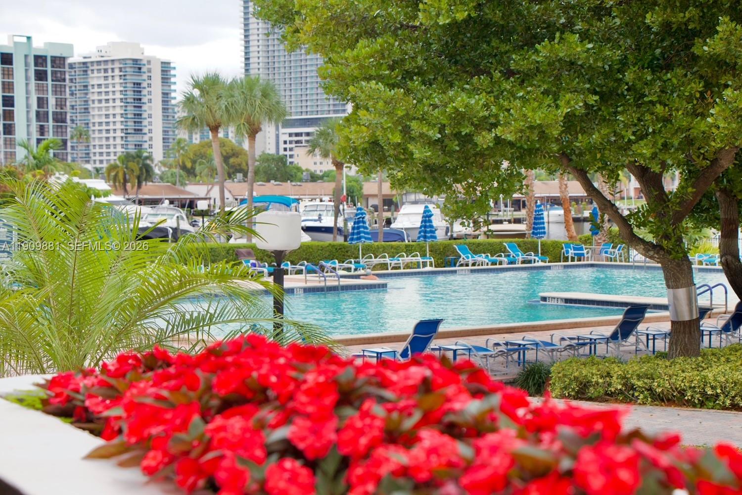 A swimming pool with lounge chairs and umbrellas is surrounded by trees and buildings — 500 Three Islands Blvd # 920, Hallandale Beach FL 33009, HALLANDALE BEACH, FL