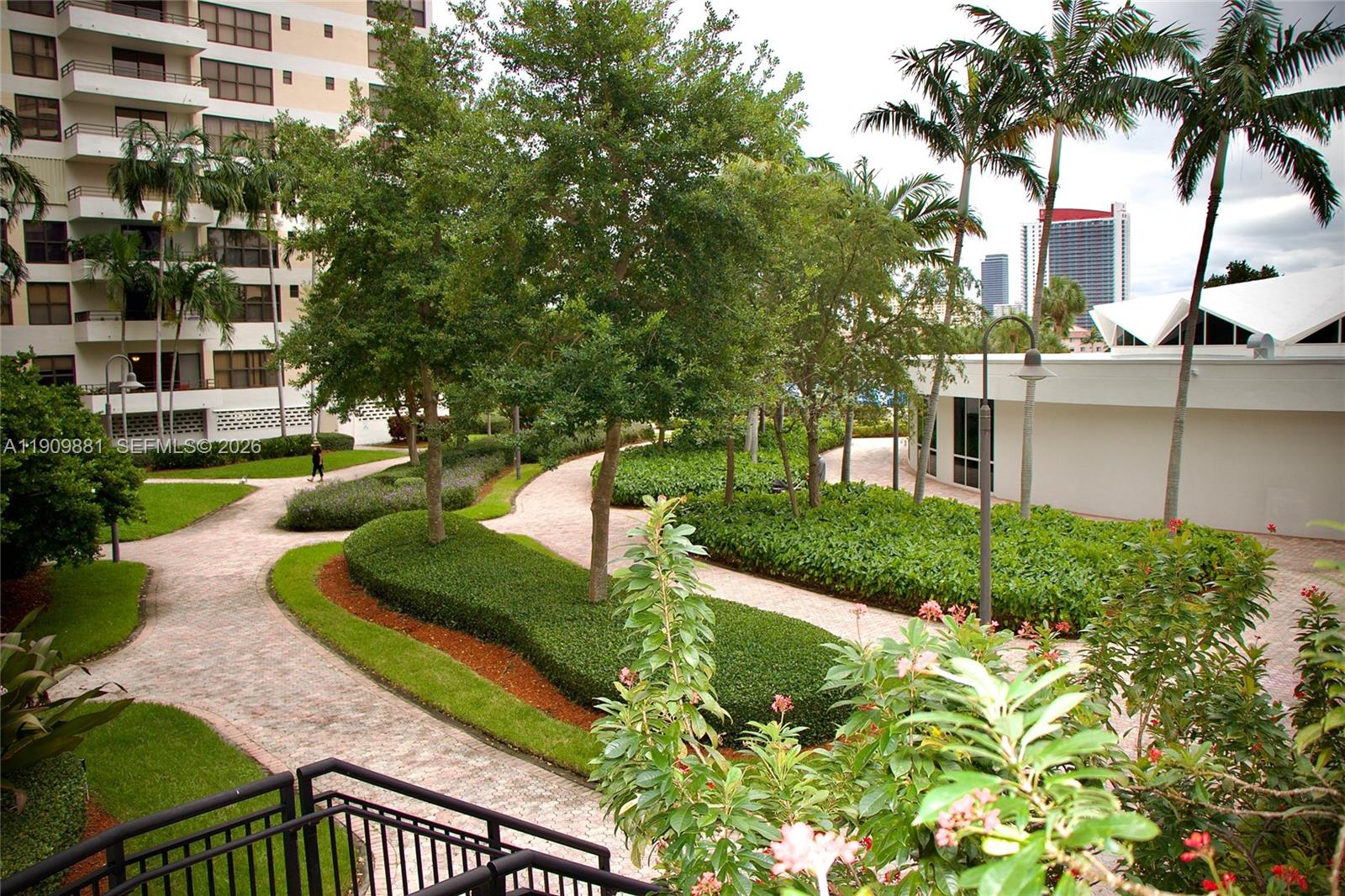 A paved walkway winds through a landscaped courtyard with trees, shrubs, and a nearby building — 500 Three Islands Blvd # 920, Hallandale Beach FL 33009, HALLANDALE BEACH, FL