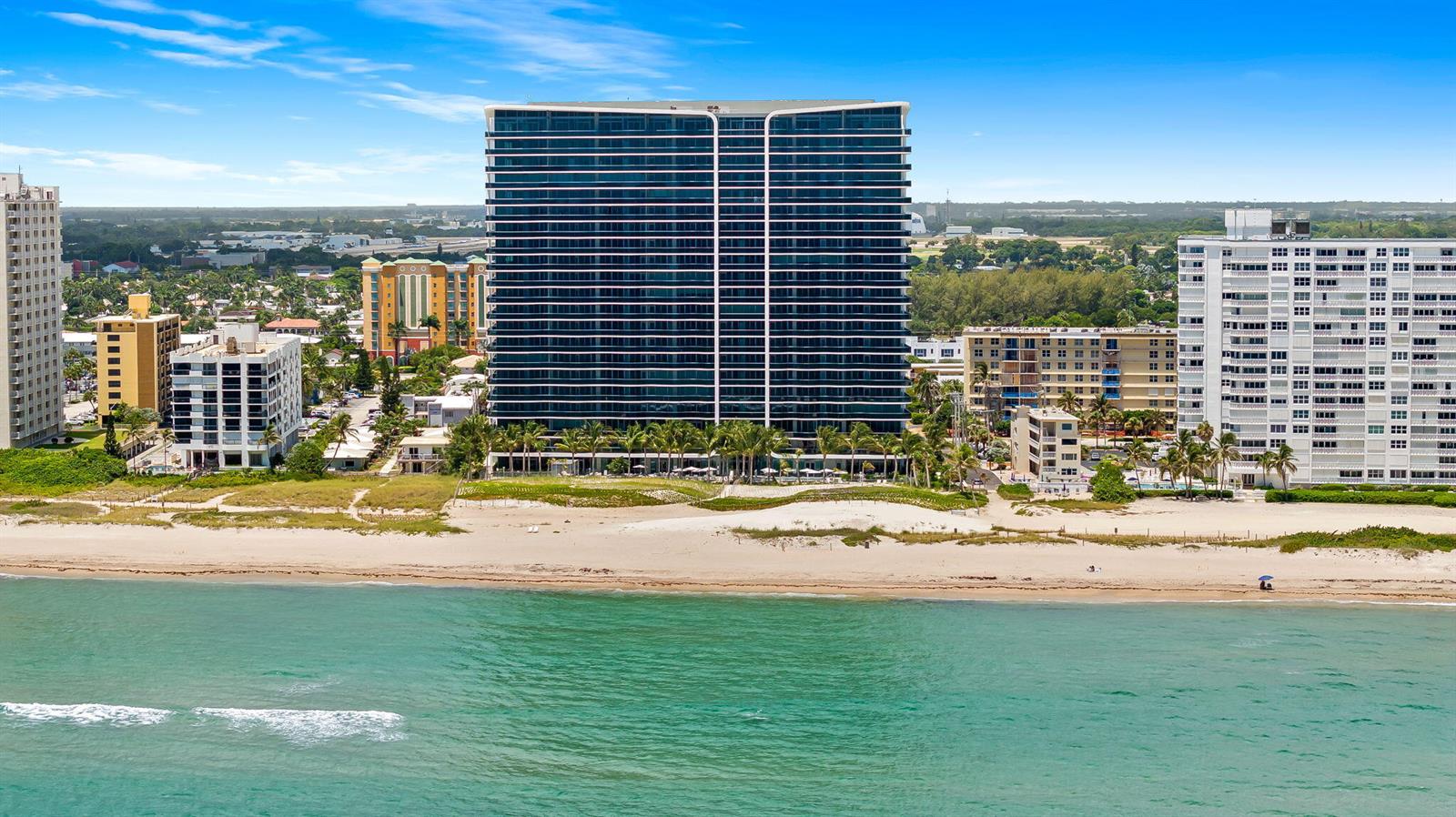 An aerial view of a tall beachfront condominium building surrounded by other coastal residential structures — 900 N Ocean Boulevard # 906, Pompano Beach FL 33062, POMPANO BEACH, FL