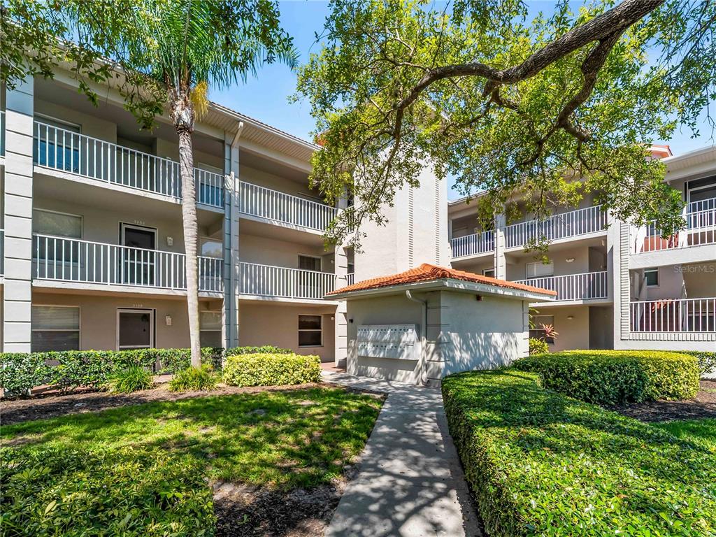 A multi-story apartment building exterior features balconies, a central mailbox kiosk, and landscaped grounds — 9330 CLUBSIDE CIRCLE UNIT 3207, SARASOTA, FL