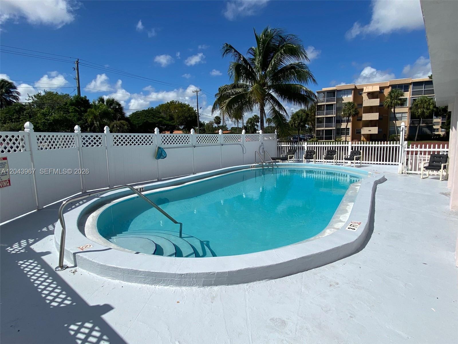 An outdoor swimming pool with a white fence and palm trees under a blue sky — 2775 Taft St # 307, Hollywood FL 33020, HOLLYWOOD, FL