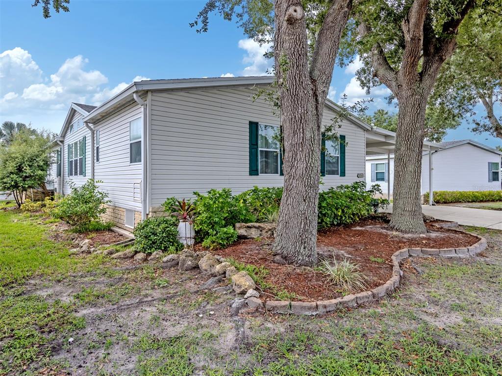 A light-colored mobile home features green shutters, a landscaped garden bed, and mature trees — 213 NATURES WAY, NORTH PORT, FL