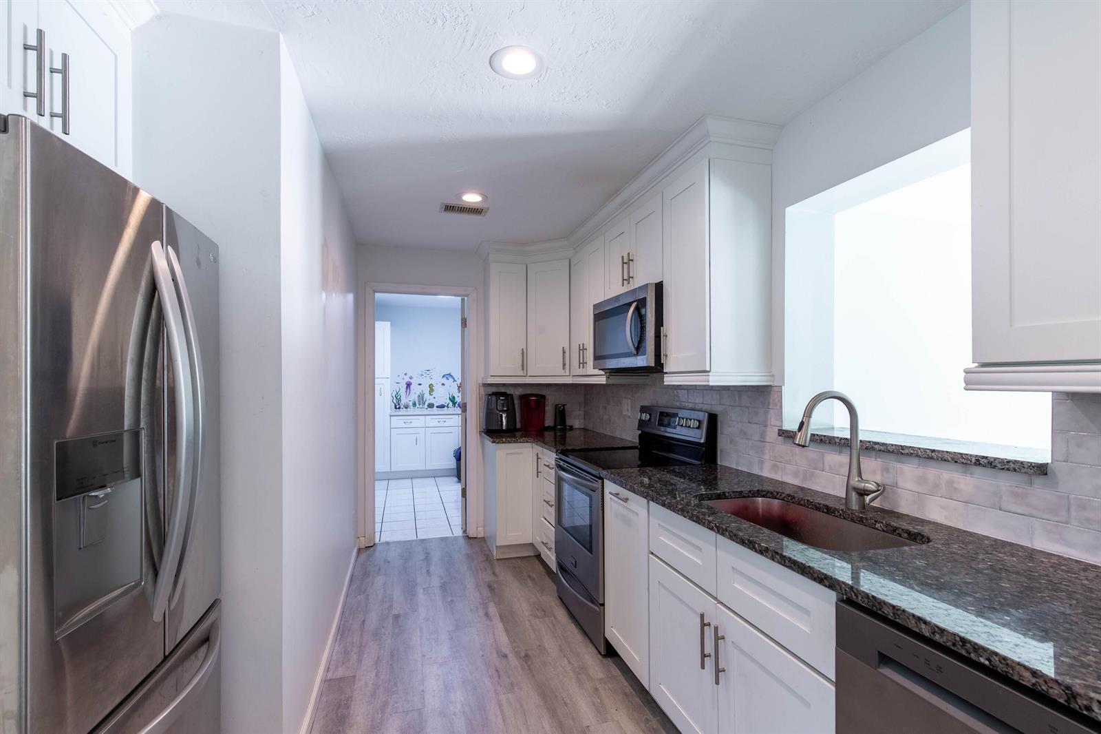 A galley kitchen features white cabinets, dark granite countertops, stainless steel appliances, and wood-look flooring — 12069 Easterly Avenue, Palm Beach Gardens FL 33410, PALM BEACH GARDENS, FL