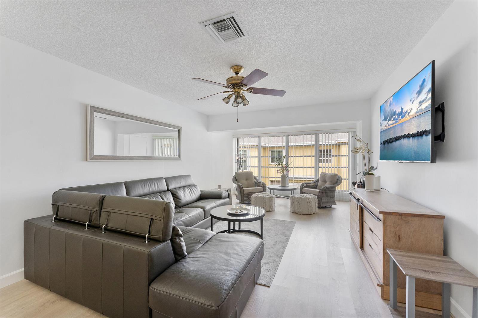 A living room features a grey sectional sofa, a television, and wicker chairs by windows — 1110 Boxwood Drive # 202, Delray Beach FL 33445, DELRAY BEACH, FL