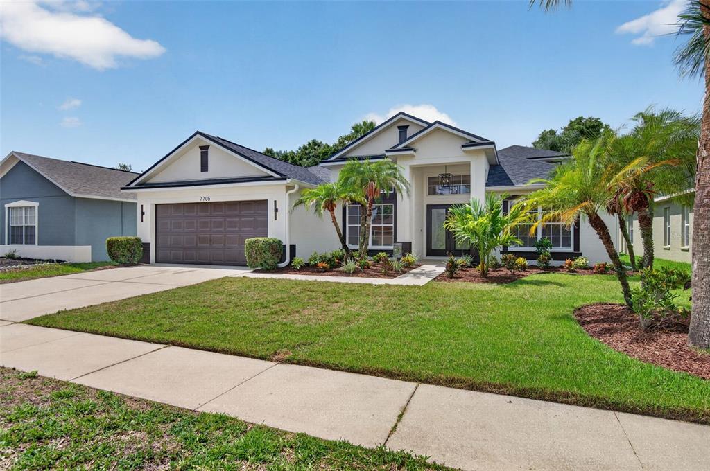 A single-story suburban house with a two-car garage, white stucco exterior, and landscaped front yard — 7705 NOTTINGHILL SKY DRIVE, APOLLO BEACH, FL