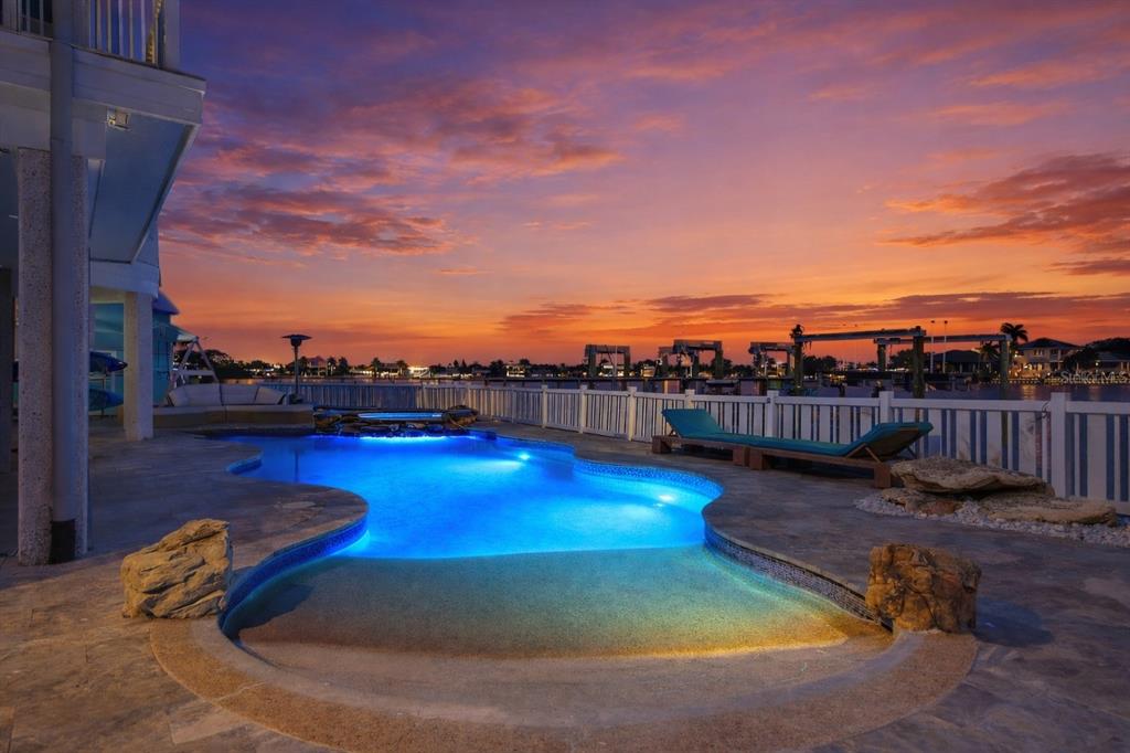 A swimming pool with blue lighting sits on a stone patio overlooking a sunset waterfront — 391 12TH AVENUE, INDIAN ROCKS BEACH, FL