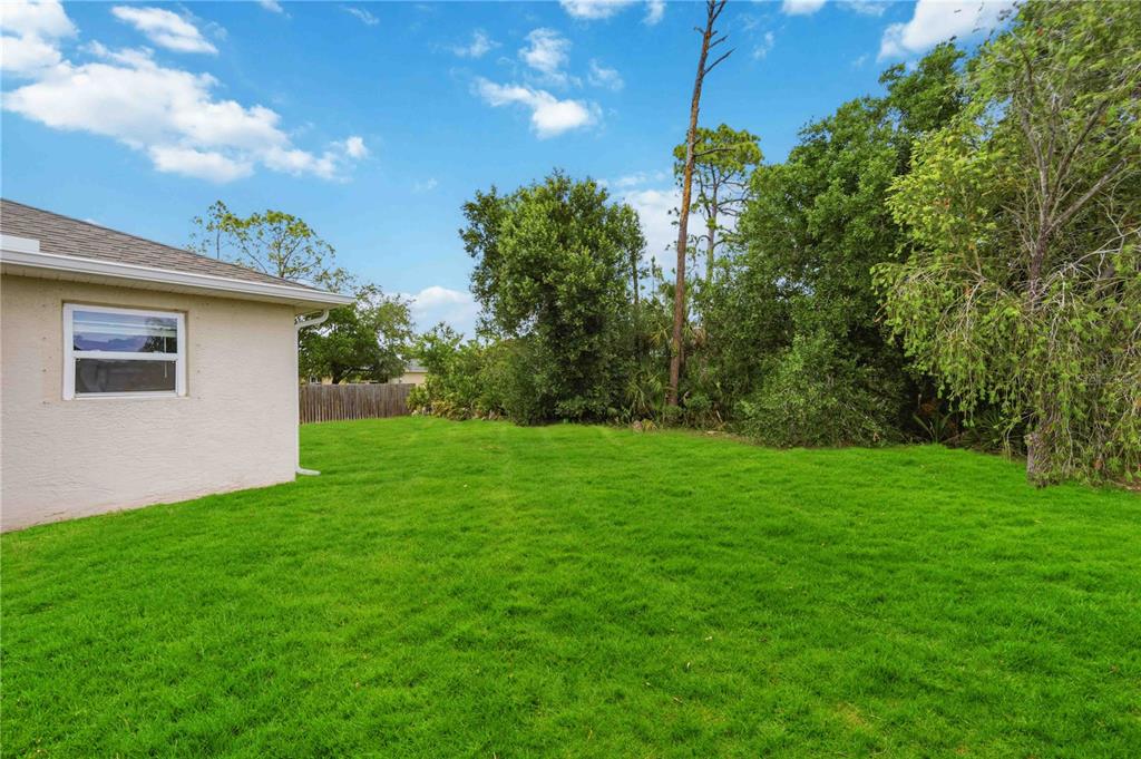 A grassy backyard view showing the side of a house and dense trees — 2260 MISTLETO LANE, NORTH PORT, FL