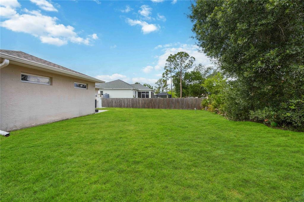 A grassy backyard features a beige house wall, a wooden fence, and mature trees — 2260 MISTLETO LANE, NORTH PORT, FL