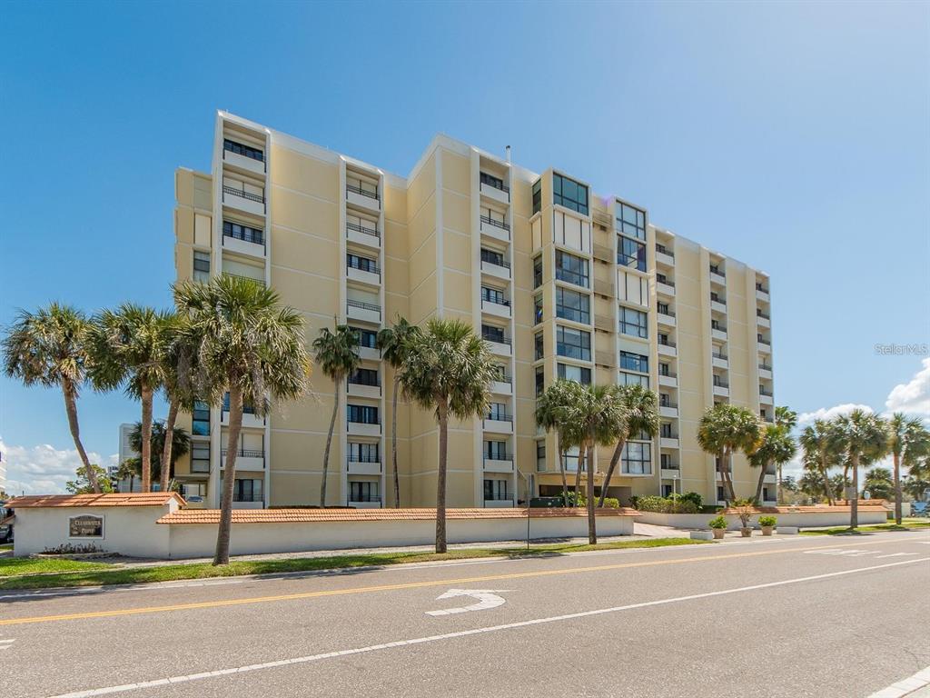 A multi-story, light-yellow residential building stands behind a low wall and palm trees — 800 S GULFVIEW BOULEVARD UNIT 305, CLEARWATER BEACH, FL