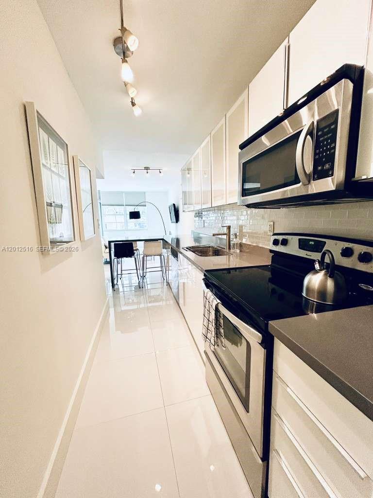 A narrow, modern galley kitchen features white cabinetry, stainless steel appliances, and glossy tile floors — 100 Lincoln Rd # 526, Miami Beach FL 33139, MIAMI BEACH, FL