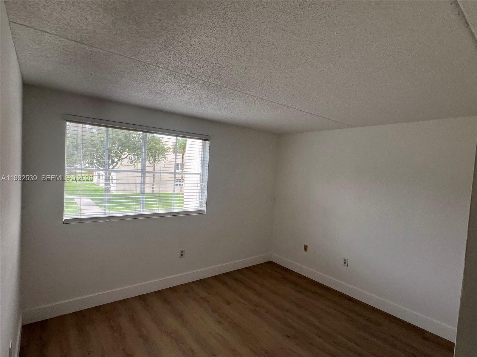 An empty room features white walls, a popcorn ceiling, and wood-look flooring with one window — 6155 NW 186th St # 207, Hialeah FL 33015, HIALEAH, FL