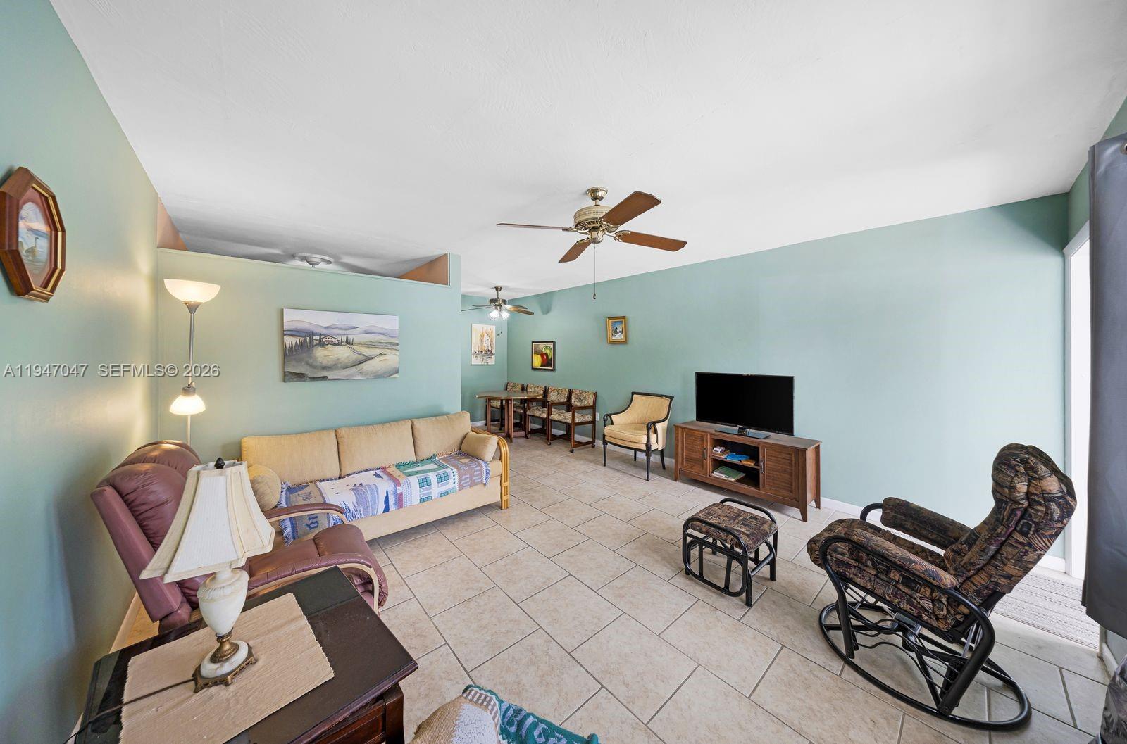 A living room with light blue walls, tile flooring, a ceiling fan, and various furniture — 15610 NE 6th Ave # 7D, Miami FL 33162, MIAMI, FL