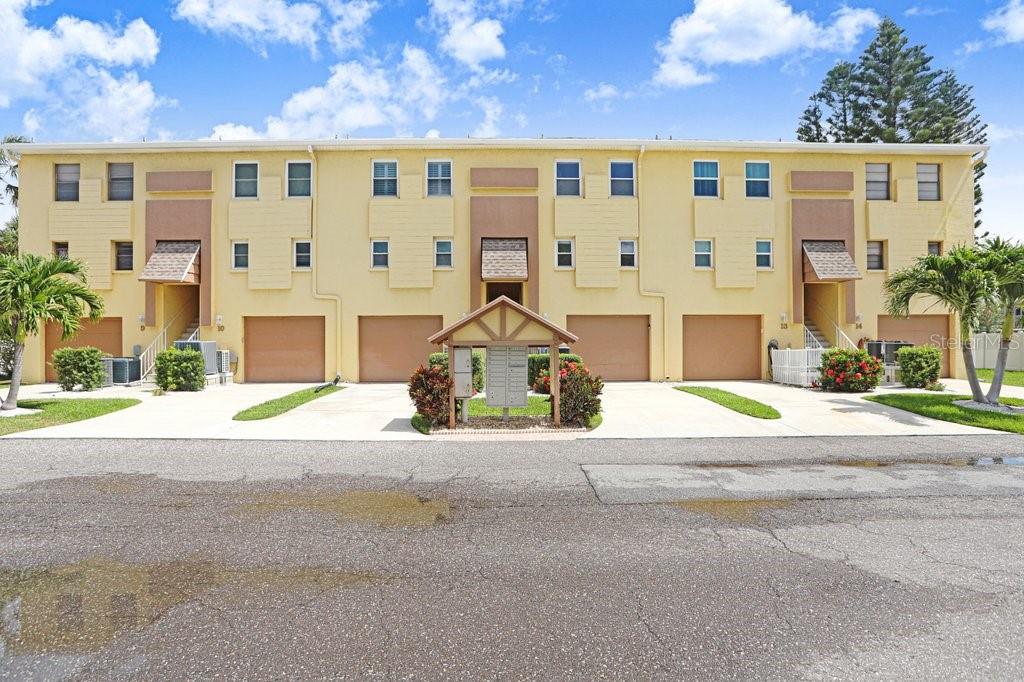 A three-story yellow townhouse complex features multiple garage doors and a central mailbox kiosk — 314 WINDRUSH BOULEVARD UNIT 12, INDIAN ROCKS BEACH, FL