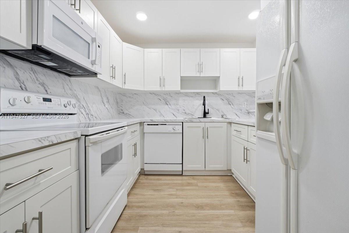 A kitchen features white cabinets, a marble-patterned backsplash, white appliances, and light wood-look flooring — 6960 Miami Gardens Dr # 124, Hialeah FL 33015, HIALEAH, FL