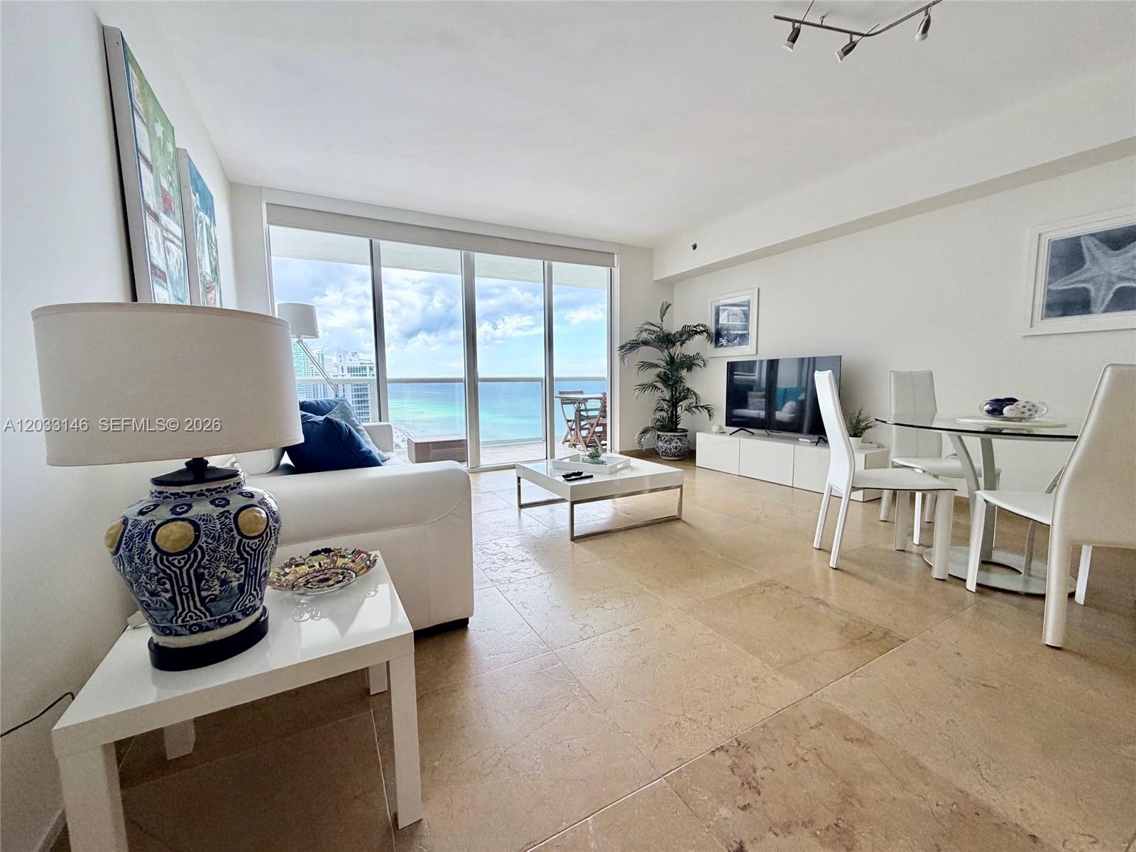 A living and dining area features tile floors, white furniture, and a balcony with ocean views — 1830 S Ocean Dr # 2412, Hallandale Beach FL 33009, HALLANDALE BEACH, FL