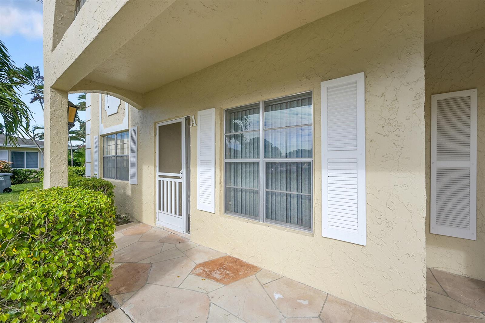 A beige stucco building exterior features a stone patio, white shutters, and a screen door — 13791 Flora Place # A, Delray Beach FL 33484, DELRAY BEACH, FL
