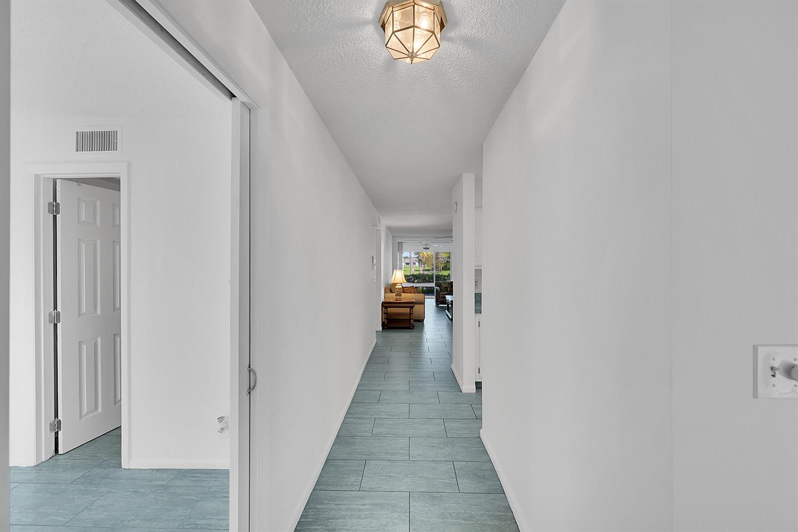 A hallway with light blue tile flooring leads toward a living area with a view — 13791 Flora Place # A, Delray Beach FL 33484, DELRAY BEACH, FL