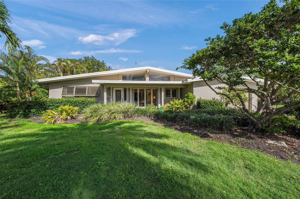 A single-story mid-century modern house with a low-pitched roof sits behind a grassy lawn — 7616 COVE TERRACE, SARASOTA, FL