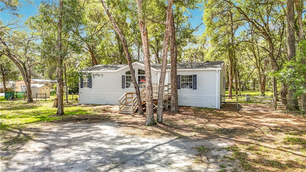 A light gray manufactured home sits in a wooded lot with a wooden front deck — 18118 PORSCHE PLACE, SPRING HILL, FL