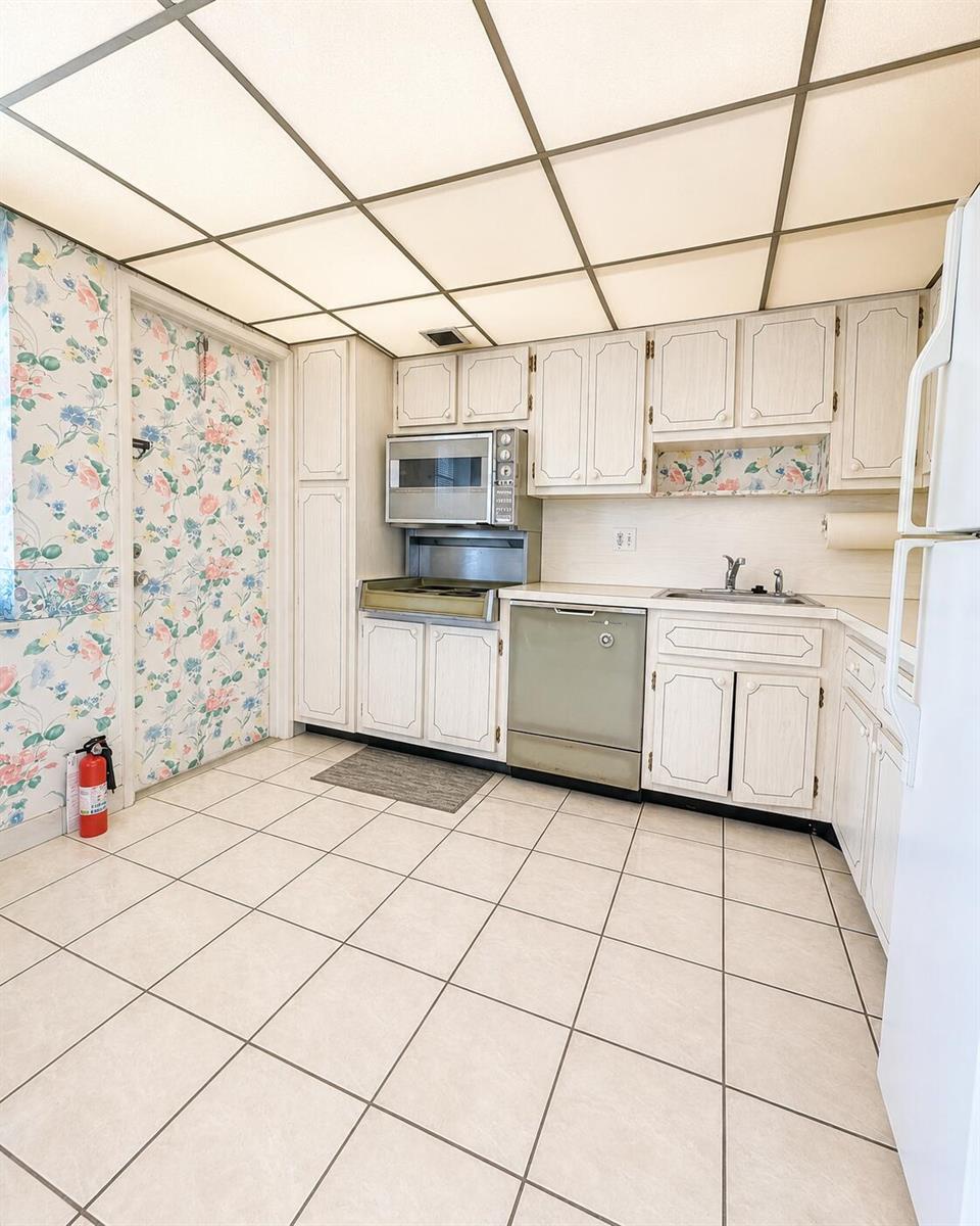 A kitchen features white cabinetry, a drop ceiling, floral wallpaper, and light-colored tile flooring — 2900 N Palm Aire Drive # 307, Pompano Beach FL 33069, POMPANO BEACH, FL