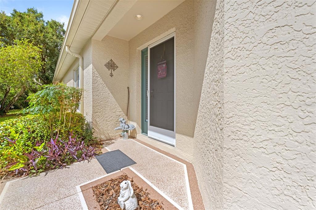 A beige stucco home exterior features a front entryway with a screen door and landscaping — 11132 HEATHROW AVENUE, SPRING HILL, FL