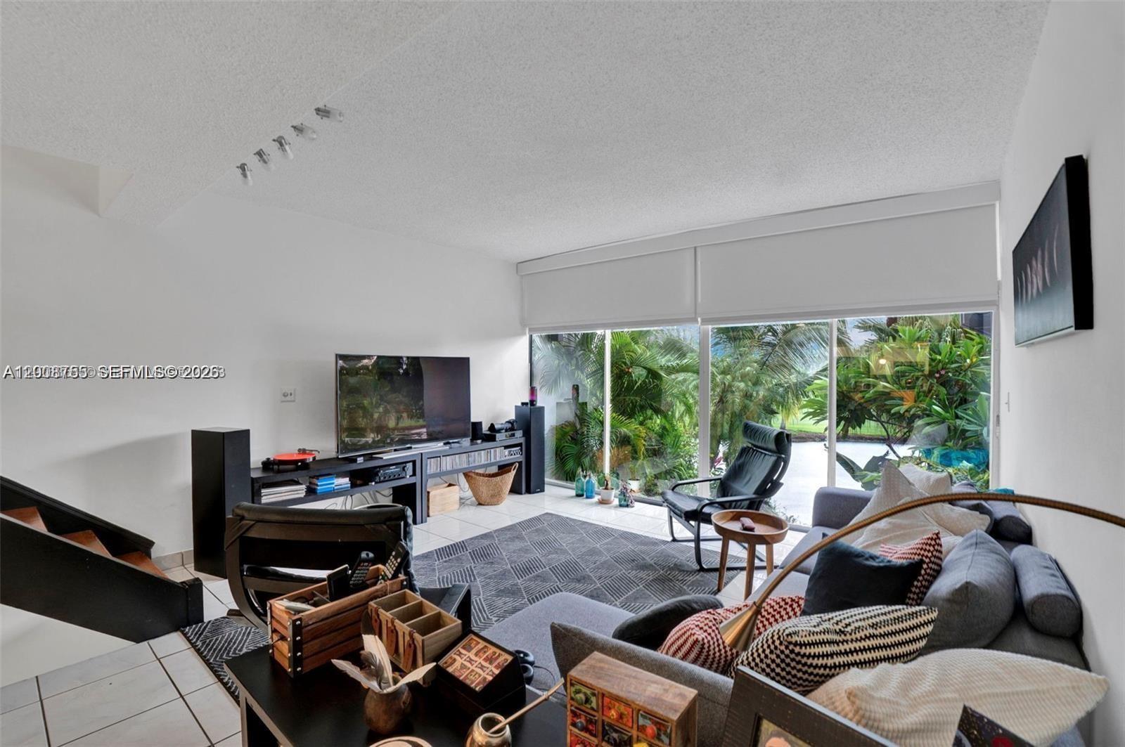 A living room features a grey sofa, a television console, and large windows overlooking palm trees — 6821 Brookline Dr # 6821, Hialeah FL 33015, HIALEAH, FL