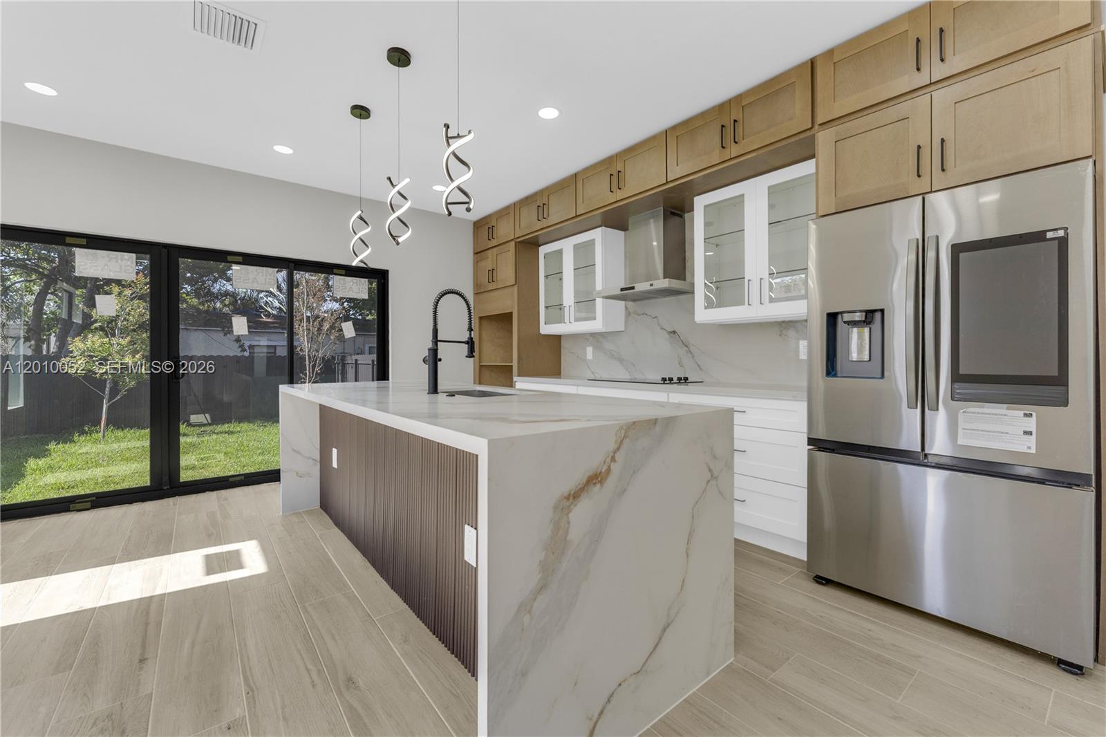A modern kitchen features a marble waterfall island, wood-toned cabinetry, and a stainless steel refrigerator — 1818 NW 49th St, Miami FL 33142, MIAMI, FL