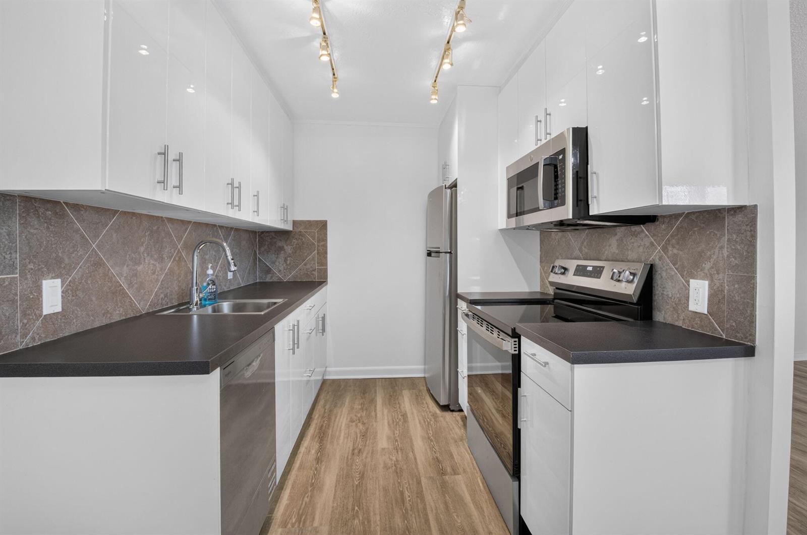 A galley kitchen features white cabinets, dark countertops, stainless steel appliances, and wood-look flooring — 3080 Cornwall D, Boca Raton FL 33434, BOCA RATON, FL