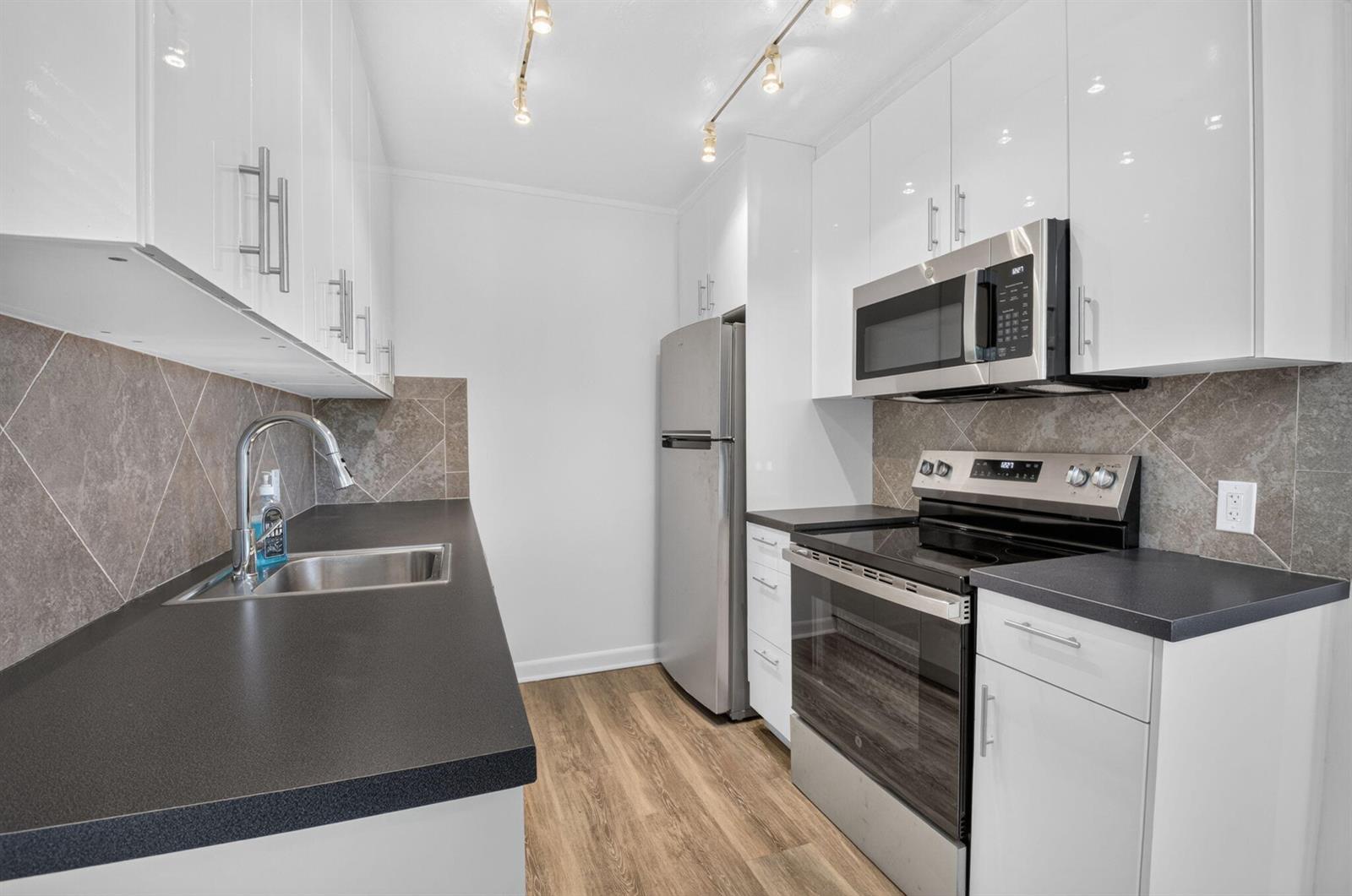 A galley kitchen features white cabinets, stainless steel appliances, dark countertops, and wood-look flooring — 3080 Cornwall D, Boca Raton FL 33434, BOCA RATON, FL