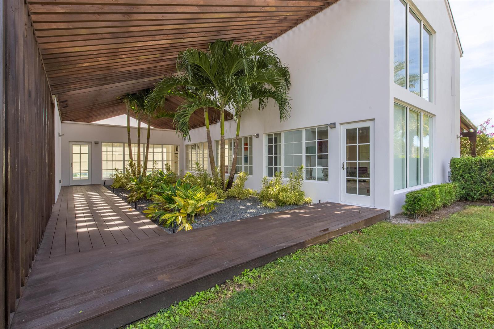 A covered wooden deck features a central garden with palm trees beside a white house — 14432 Rolling Rock  (house) Place, Wellington FL 33414, WELLINGTON, FL