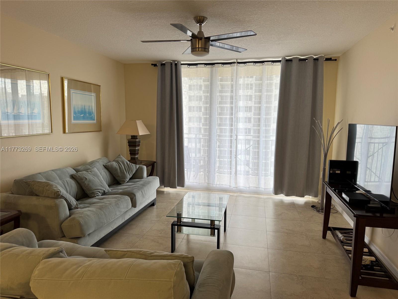 A living room features a light-colored sofa, glass coffee table, ceiling fan, and balcony curtains — 2080 S Ocean Dr # 1405, Hallandale Beach FL 33009, HALLANDALE BEACH, FL