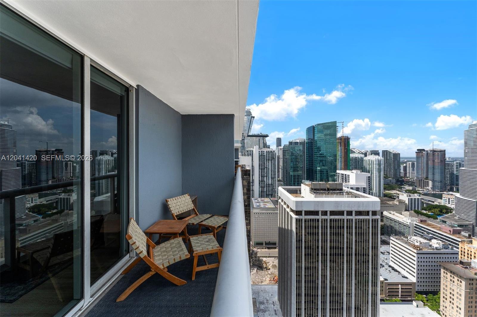 A high-rise balcony features two wooden chairs with ottomans and a view of city skyscrapers — 50 Biscayne Blvd # 4603, Miami FL 33132, MIAMI, FL