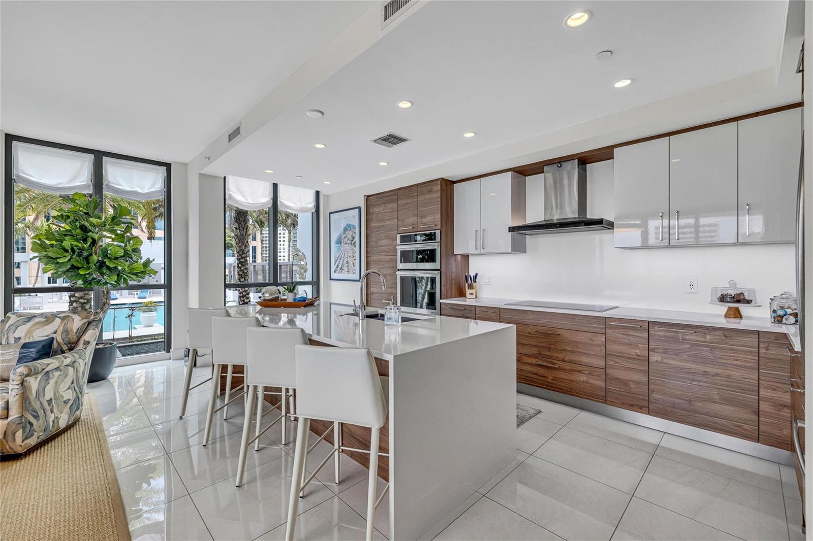 A modern kitchen features a large white island, wood cabinetry, and floor-to-ceiling windows — 100 E Las Olas Boulevard # 1701, Fort Lauderdale FL 33301, FORT LAUDERDALE, FL
