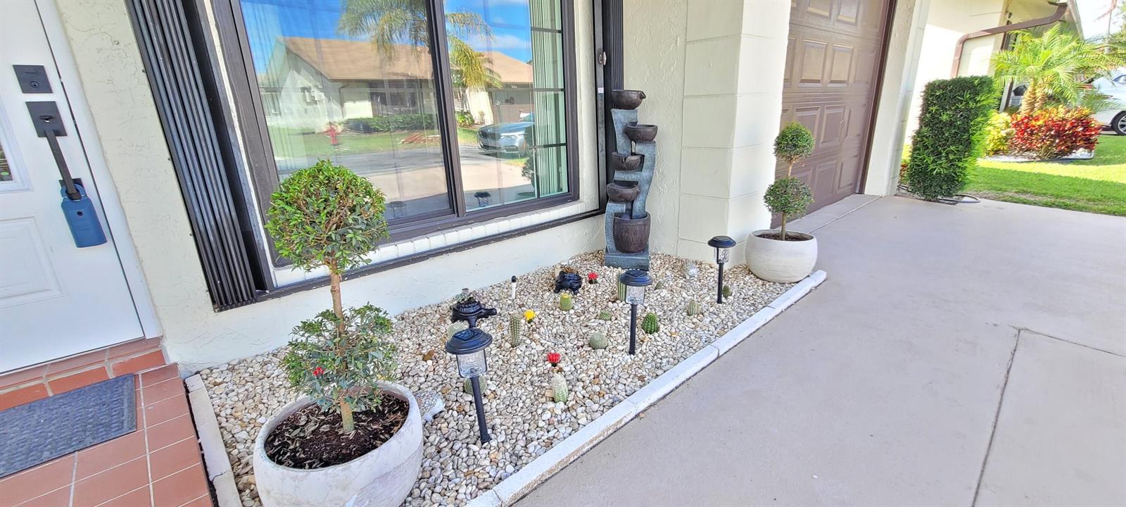 A small garden bed with gravel, potted topiary trees, and cacti sits beside a house exterior — 6209 Olivewood Circle, FL 33463, FL