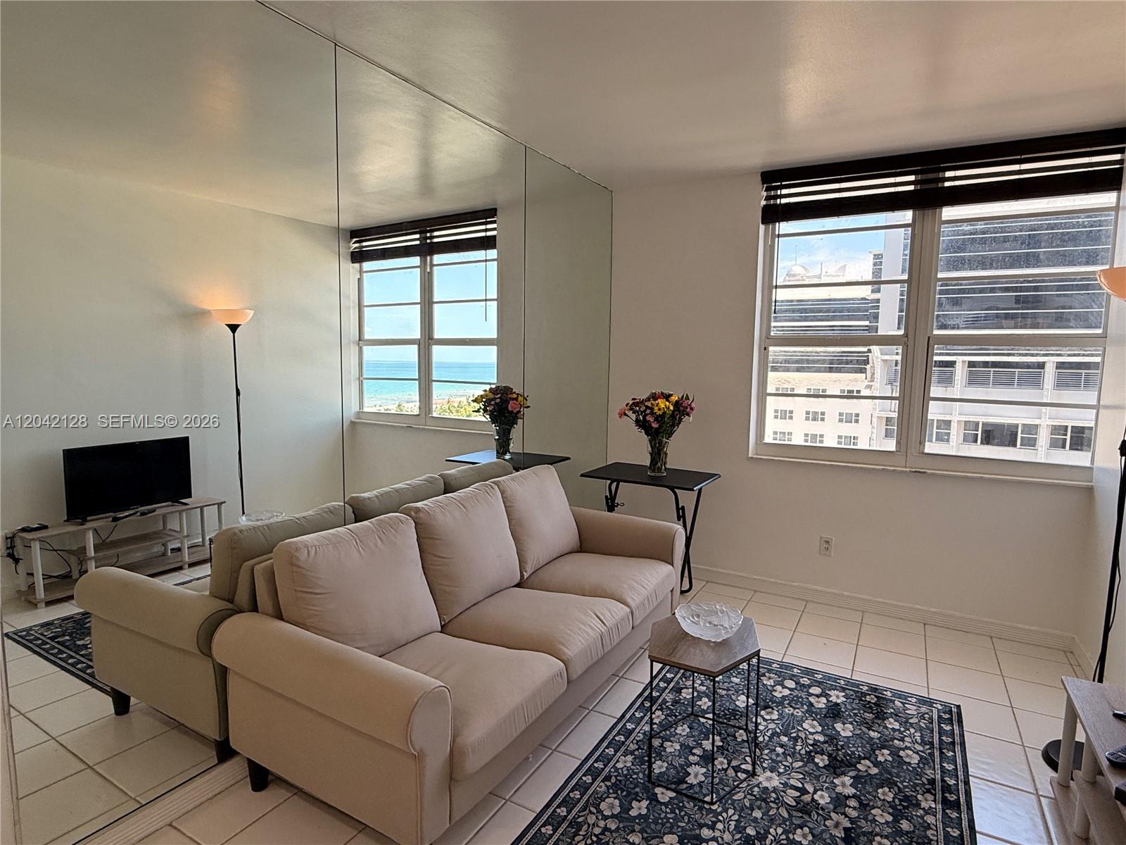 A living room features a beige sofa, tiled floors, a mirrored wall, and ocean views — 100 Lincoln Rd # 1025, Miami Beach FL 33139, MIAMI BEACH, FL
