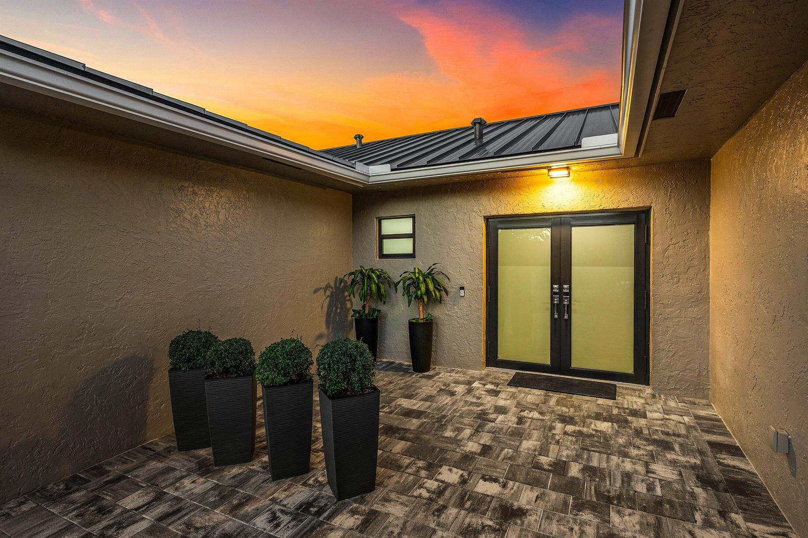 A paved courtyard features potted plants, a double glass door, and a sunset sky overhead — 21212 Escondido Way, Boca Raton FL 33433, BOCA RATON, FL