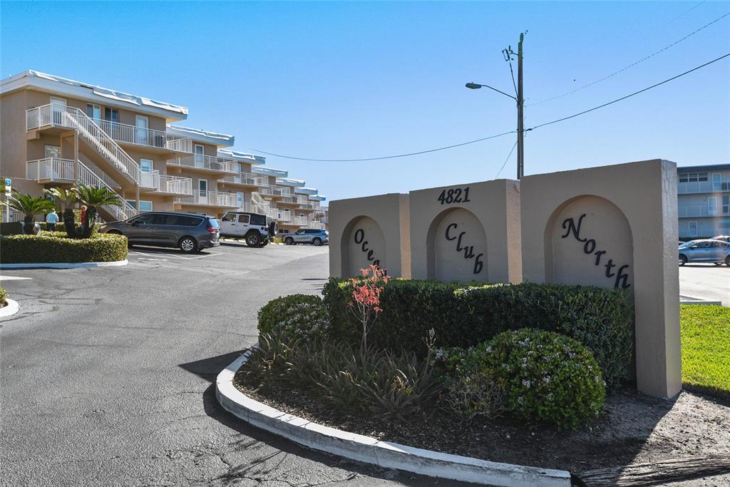 The exterior of a multi-story apartment complex features a prominent sign reading Ocean Club North — 4821 SAXON DRIVE UNIT B-213, NEW SMYRNA BEACH, FL