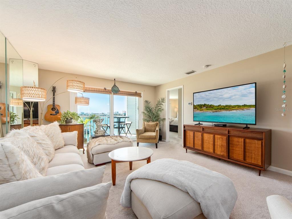A living room features a sofa, ottomans, a television on a wooden console, and a balcony — 610 ISLAND WAY UNIT 407, CLEARWATER BEACH, FL