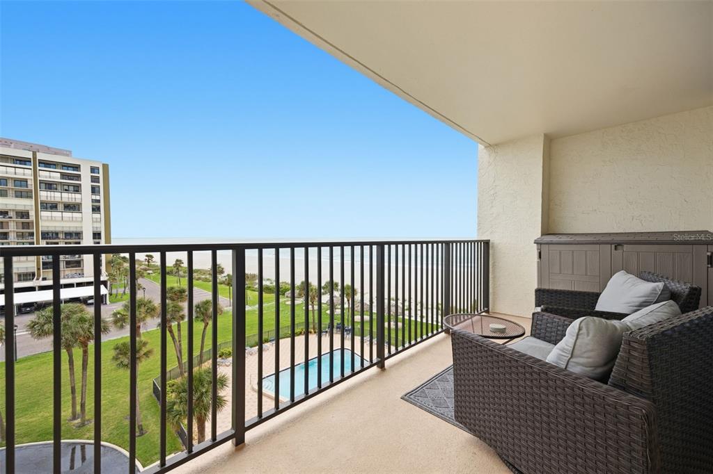 A balcony with wicker furniture overlooks a swimming pool, green lawn, and the ocean — 1460 GULF BOULEVARD UNIT 501, CLEARWATER, FL