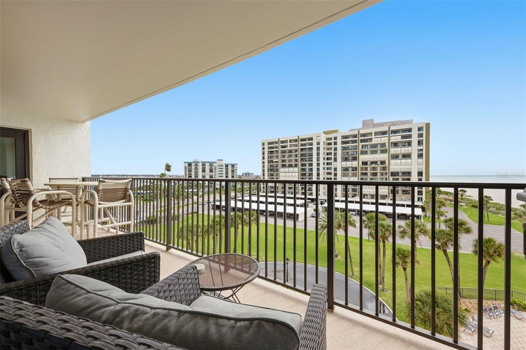 A balcony with wicker furniture overlooks a grassy lawn, palm trees, and a large building — 1460 GULF BOULEVARD UNIT 501, CLEARWATER, FL