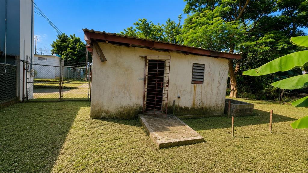 A small, weathered concrete outbuilding with a metal security door stands in a grassy yard — CO Villa Caridad AMPARO ST. UNIT D-10, CAROLINA, PR