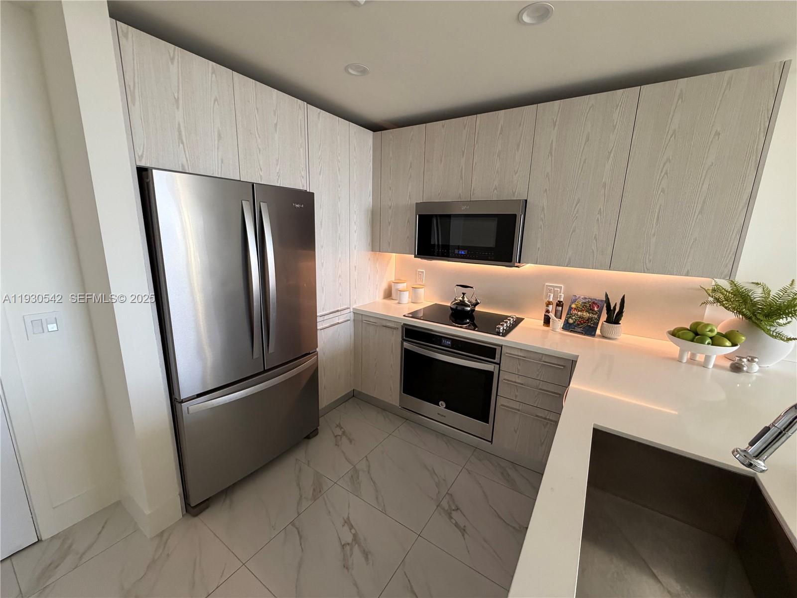 A modern kitchen features light wood-grain cabinets, stainless steel appliances, and white marble-patterned tile flooring — 2000 Metropica Way # 2108, Sunrise FL 33323, SUNRISE, FL