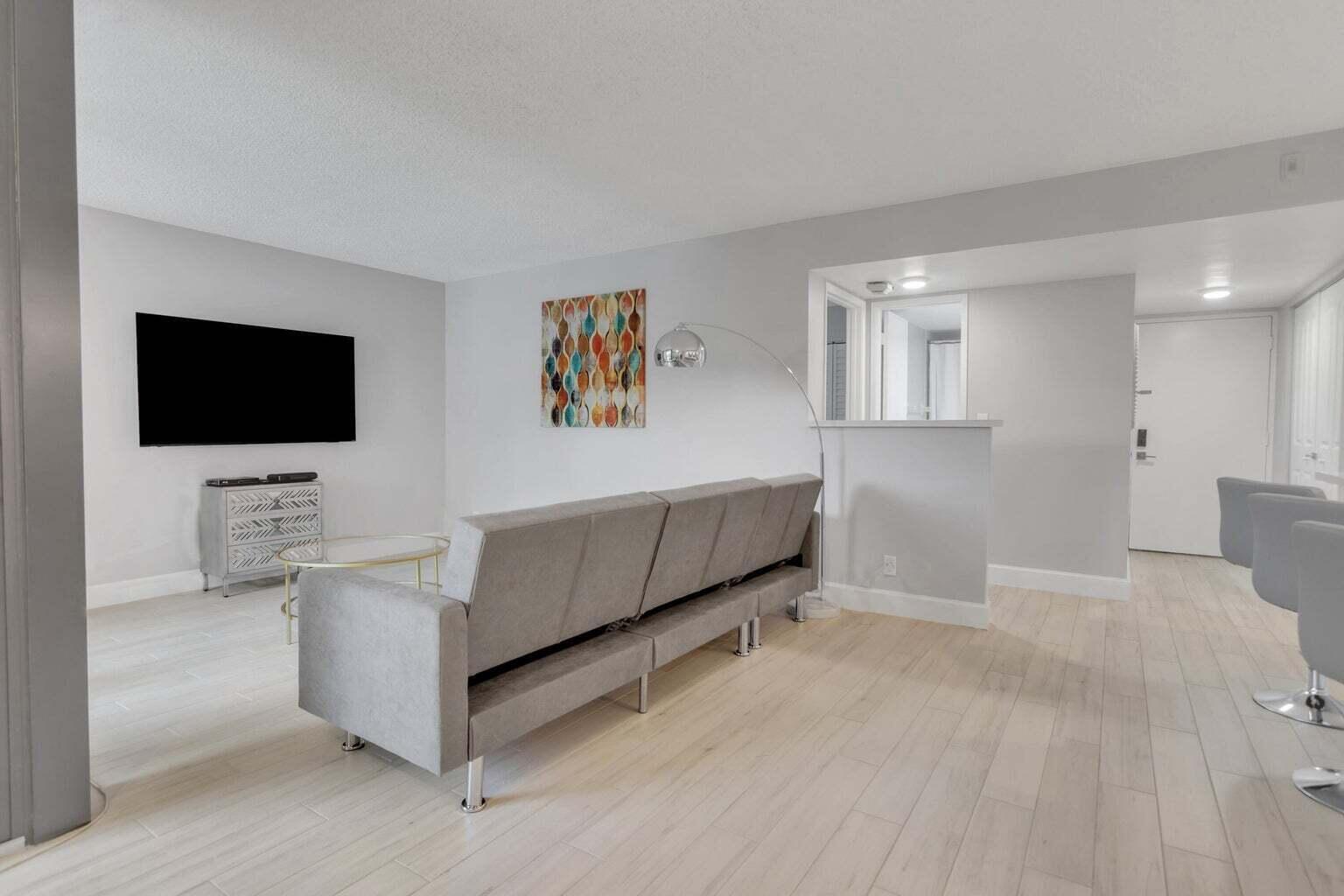 A living room features light wood-look flooring, a gray sofa, a wall-mounted television, and artwork — 955 Dotterel Road, Delray Beach FL 33444, DELRAY BEACH, FL