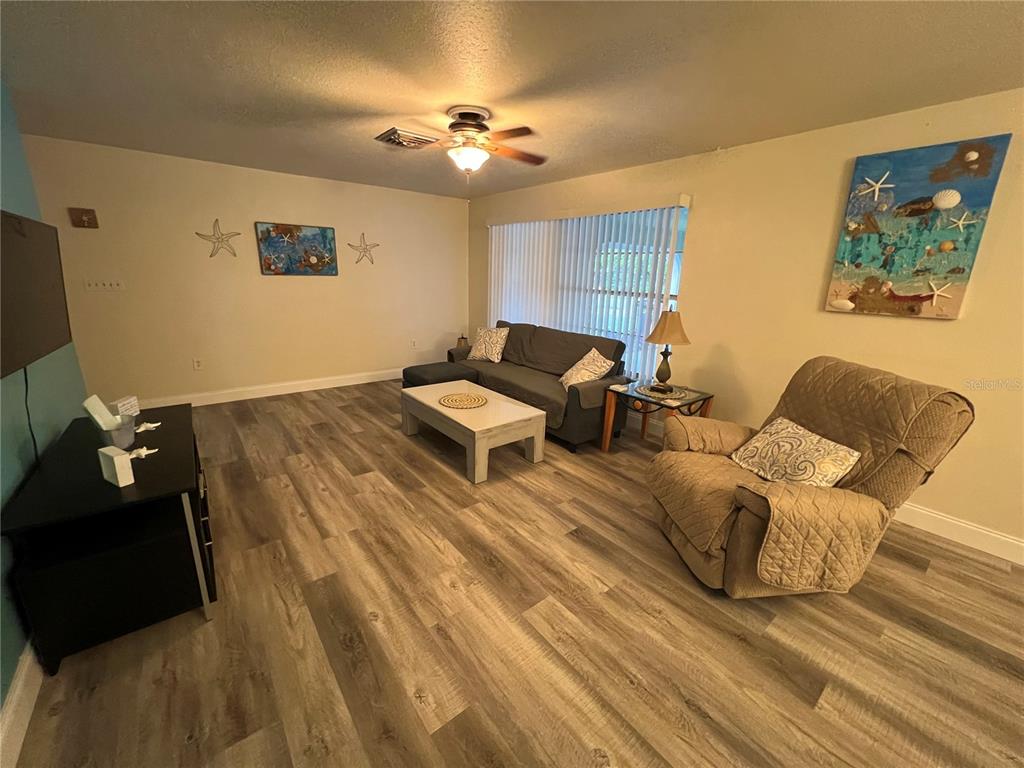 A living room features wood-look flooring, a gray sofa, a tan recliner, and coastal wall decor — 1406 N SATURN AVENUE, CLEARWATER, FL