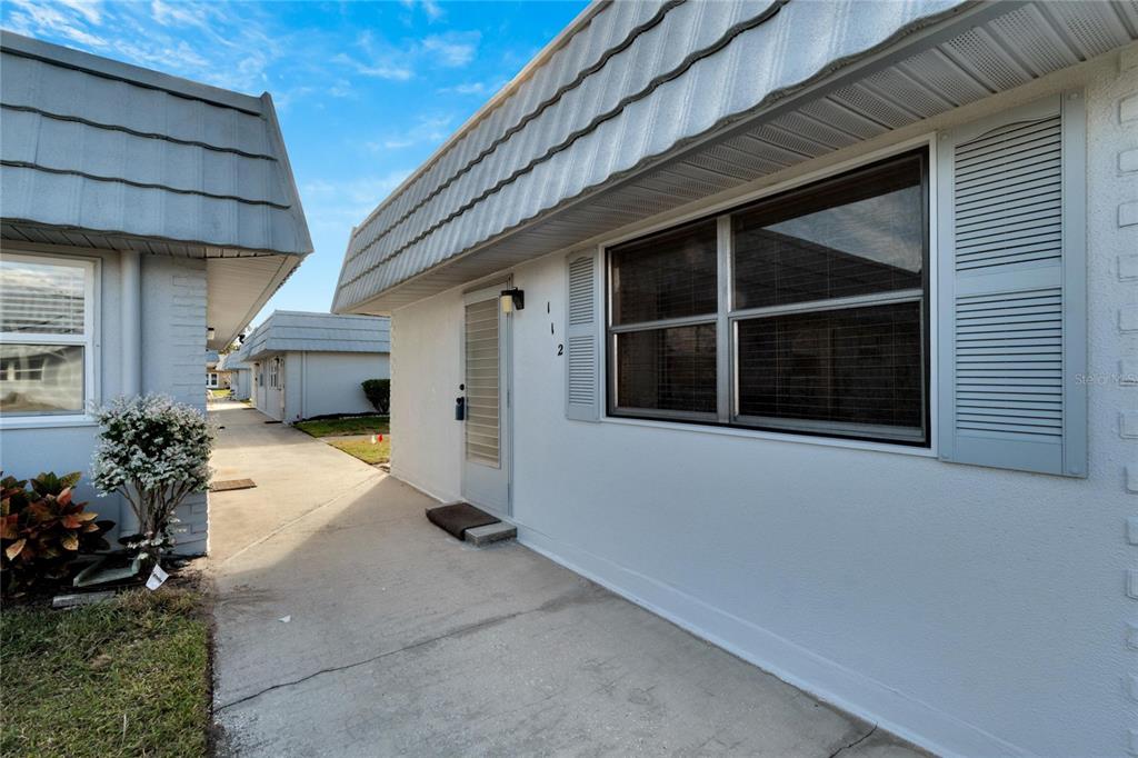 A white exterior wall with a front door, a window with shutters, and a concrete walkway — 202 BEDFORD TRAIL UNIT 112, SUN CITY CENTER, FL